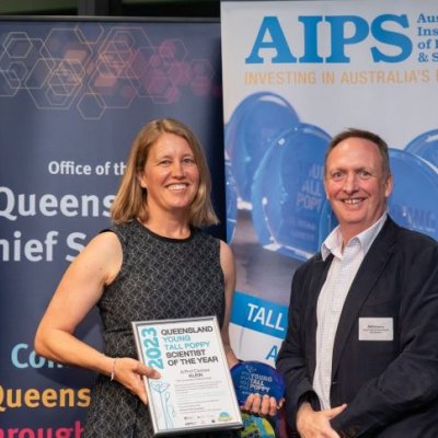 Two people stand smiling in front of banners at an awards night, one of them is holding an award.